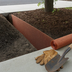 Copper mesh root barrier standing upright in an open trench between a tree bed and a concrete sidewalk, with a roll of copper screen beside it