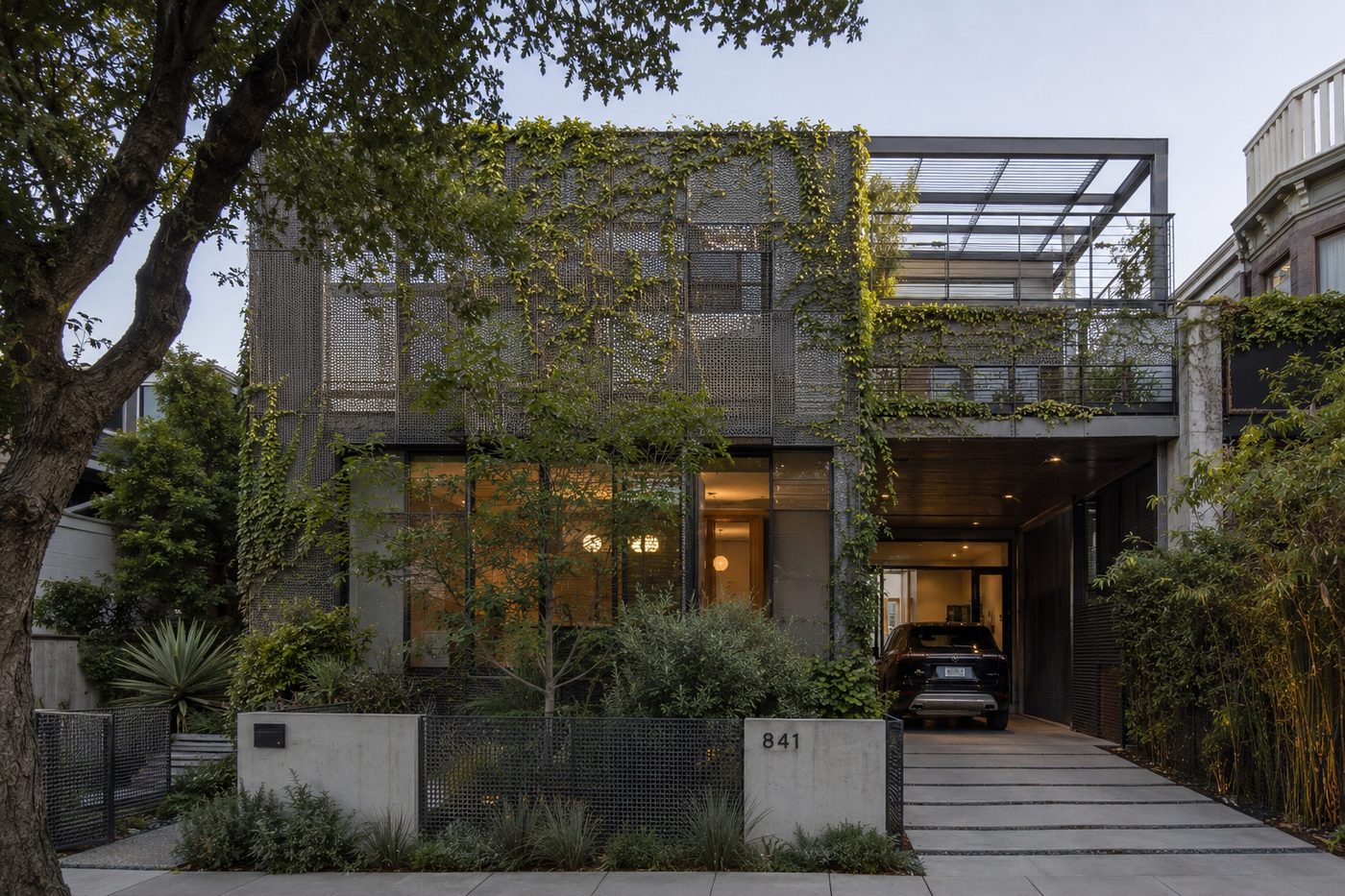 Perforated metal facade on a modern home, softened by climbing vines, with a woven mesh garden fence at the street
